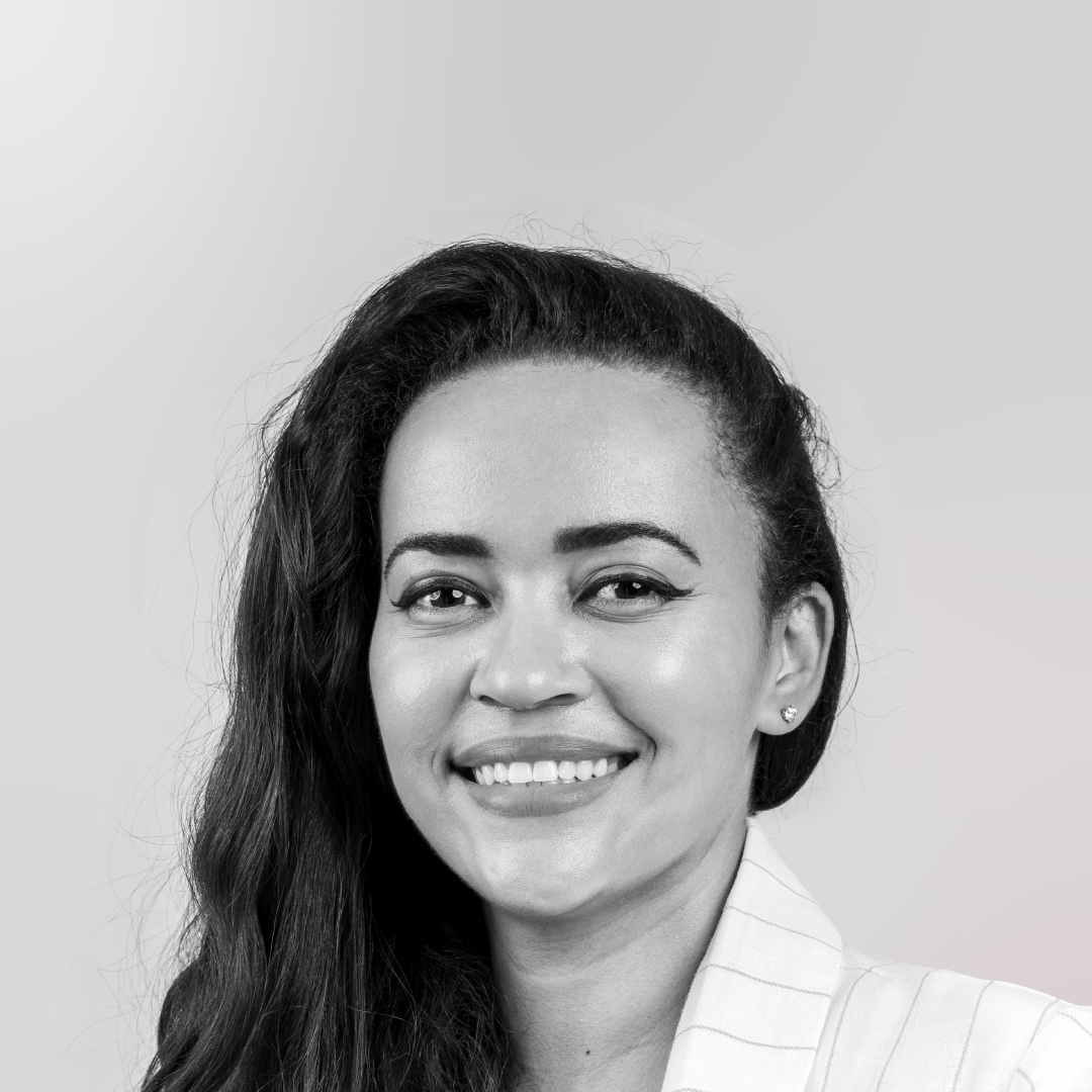 Black-and-white headshot of Aminah Moore smiling, with long hair swept to one side, wearing a light pinstriped blazer against a soft grey background.